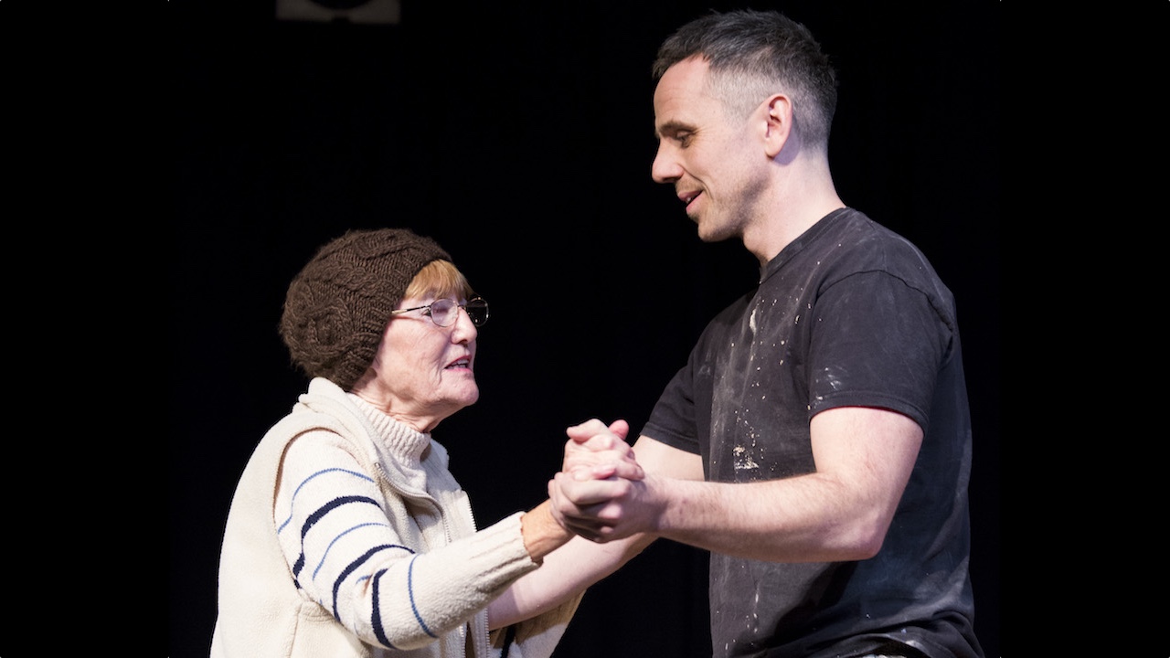 A tall middle aged man in a black t-shirt, speckled with white paint, dances with an elderly woman wearing a brown wooly skull cap and a light beige wooly cardigan.