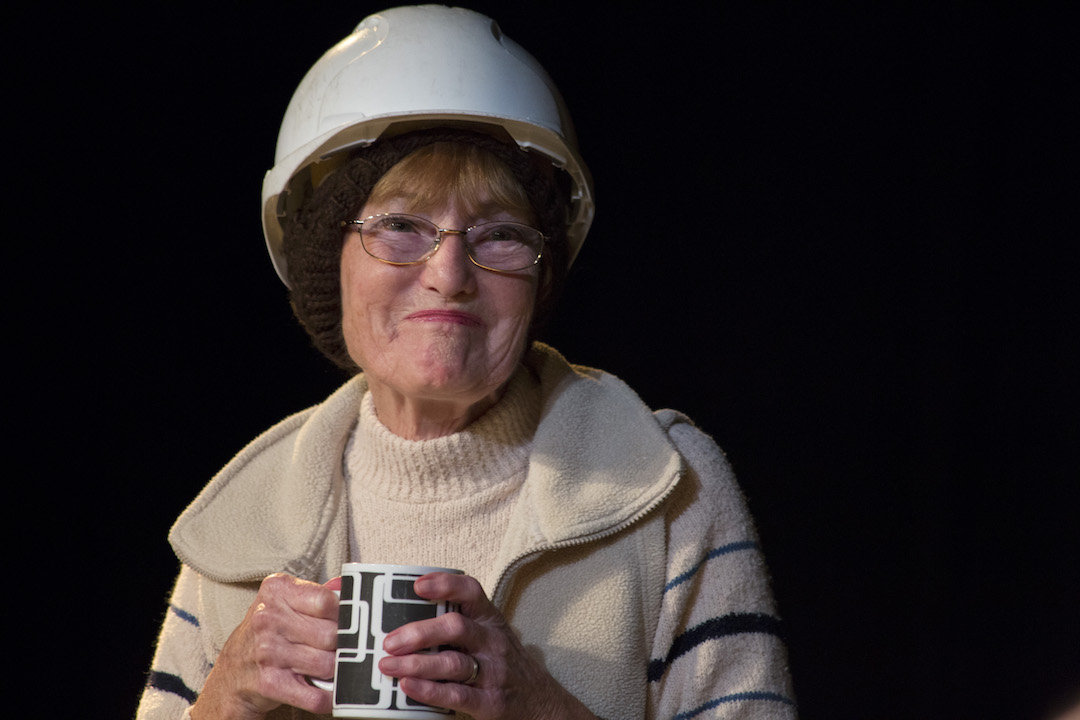 An elderly lady, with spectacles, smiles at the camera, with a cup of tea clenched at her chest. She is wearing a white hard hat over a brown wooly skull cap and a light beige wooly jumper under a beige fleece body warmer.