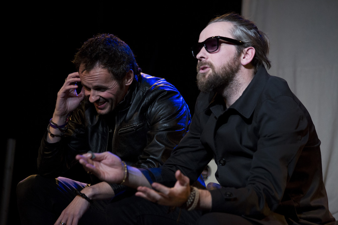 Two men in black clothing sit next to each other. The man on the left and towards the back of the picture is on his phone, gimmacing and looking at the floor. The man on the right and nearer the front of the picture has a full beard and is wearing sunglasses. He has his hands out in front of him, palms up, as though explaining or pleading with someone stood over him out of shot.