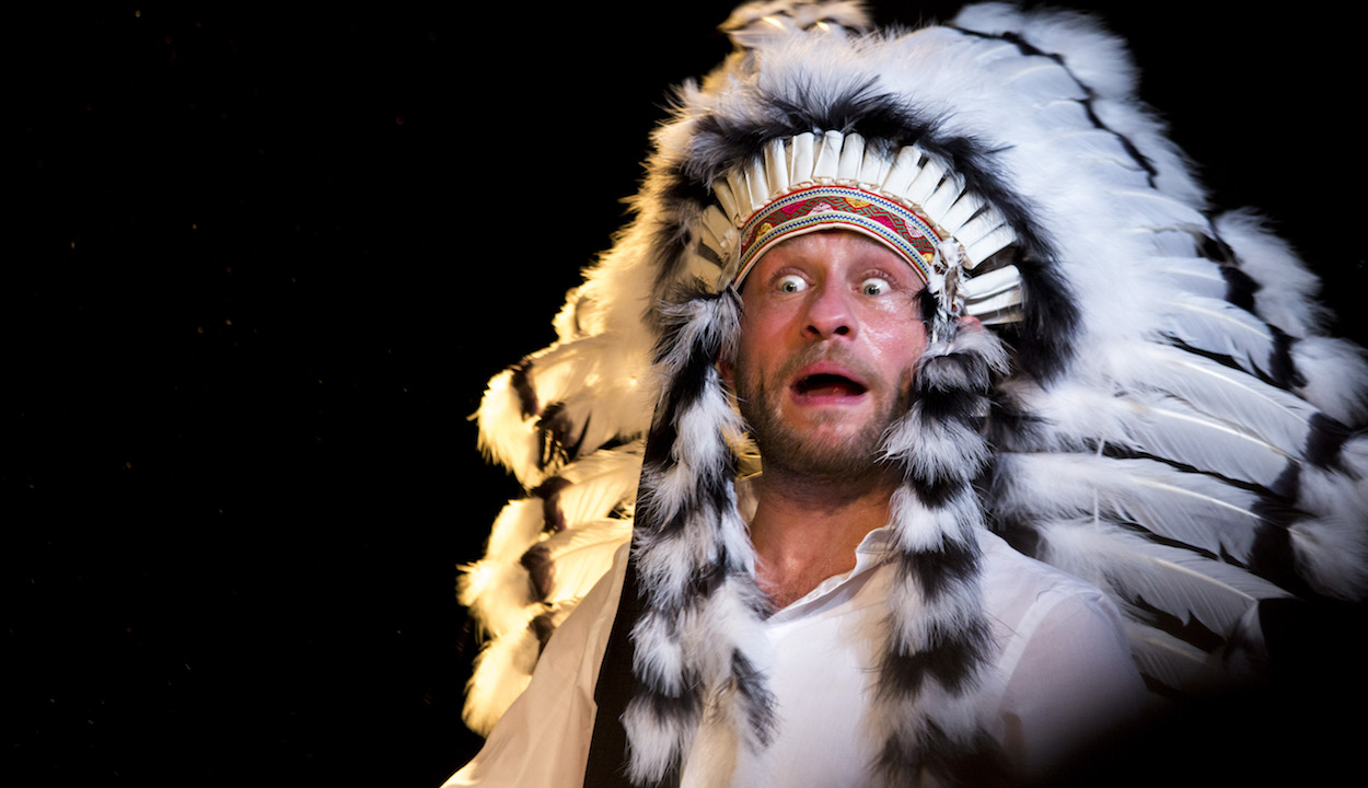 A white man wearing traditional Native American headgear has a strange expression on his face, with his mouth open and his eyes wide open.