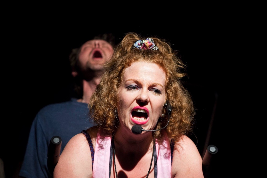 At the forefront of the image is a woman seemingly shouting into a headset microphone. Out of focus behind her is a man appearing to simulate a sexual act on the woman.